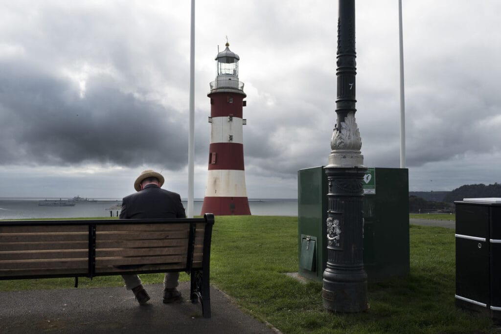 A street photo of an elderly man on a bench with a lighthouse in the background, by Ryan Hardman